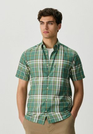 Young man wearing a green, white, and yellow plaid short-sleeve button-up shirt with beige pants, standing with hands in pockets.