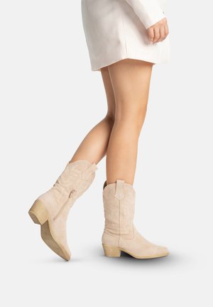 Piernas con botas vaqueras estilo occidental hasta la mitad de la pantorrilla de ante beige claro con detalles bordados y tacones de bloque de madera, combinadas con una falda corta blanca.
