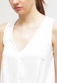 Top bianco senza maniche con scollatura a V profonda, texture di raso liscio e sottili dettagli di cucitura lungo le cuciture.