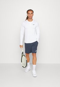 Biała, długosleeve'owa koszulka sportowa z połową zamka i czarnym logo, w zestawieniu z ciemnoszarymi szortami. Model trzyma rakietę tenisową i nosi białe sneakersy.