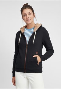 Black zip-up hoodie with a tan fleece-lined hood, contrasting drawstrings, and triangular accent details on pockets. Striped inner shirt visible.
