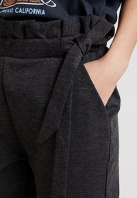 Detailní záběr na ruku osoby v kapse tmavě šedých kalhot s látkovým opaskem uvázaným v pase, která má na sobě černé tričko zastrčené dovnitř.