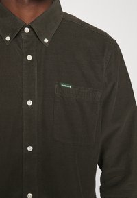 Donkergroen corduroy overhemd met knopen, voorzien van een borstzak, witte knopen en een klein groen Barbour-logo op de zak.