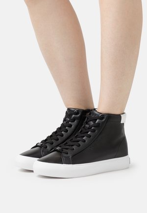 Jalat, joissa on mustat korkeavartiset tennarit valkoisilla pohjilla ja mustilla nauhoilla, seisomassa valkoisella pinnalla.