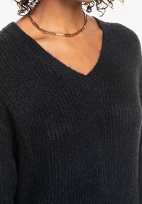 Schwarzer Strickpullover mit V-Ausschnitt, strukturierter Stoff und gerippten Kanten. Kombiniert mit einer goldenen Gliederkette.
