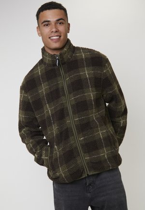 Joven sonriendo, vestido con una chaqueta de forro polar a cuadros verde y marrón oscuro con cremallera y bolsillos, de pie contra un fondo claro y liso.