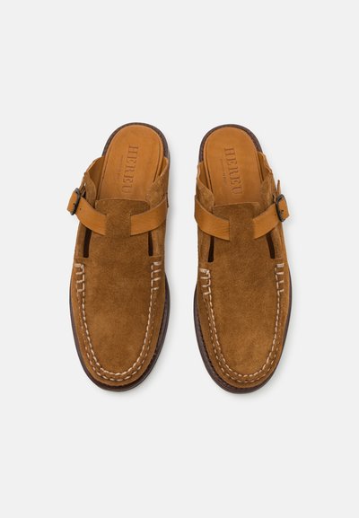 Scarpe slip-on in suede tan con punta arrotondata, dettagli cuciti e una fascia regolabile. La suola presenta una finitura marrone a contrasto.