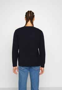 Pull en maille bleu marine foncé avec un col rond, des poignets et un ourlet côtelés, associé à un jean bleu clair. Coiffure tressée visible de dos.