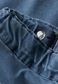 Džinsa auduma tuvplāns ar ribveida faktūru, tumši zilā krāsā un skaidru pogu. Redzamas šūšanas līnijas un elastīga jostasvietas detaļa.