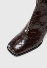 Botín de cuero marrón oscuro con estampado de cocodrilo, punta cuadrada y costura visible a lo largo de la costura frontal.