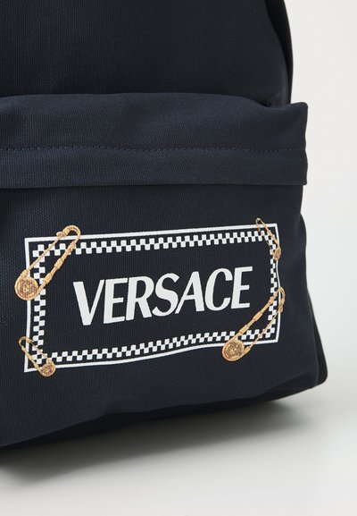 Sac à dos en toile bleu marine avec un logo "VERSACE" blanc et des épingles de sécurité dorées, doté d'un détail à bordure à carreaux sur la poche avant.