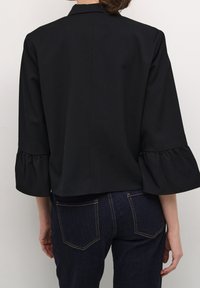 Svart kort jacka med utsvängda trekvartsärmar, slät tyg, subtila axelsömmar och en klassisk krage. Mörka denimjeans synliga.