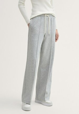 Personne portant un pantalon de survêtement gris ample avec des coutures à l'avant et une taille à cordon, associé à des baskets blanches et un haut blanc côtelé à manches longues.