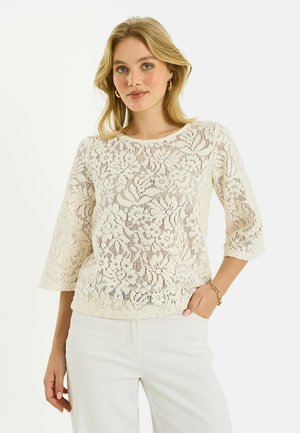 Femme portant un haut en dentelle florale crème à manches longues avec un pantalon blanc, posant avec une main dans la poche sur un fond blanc.