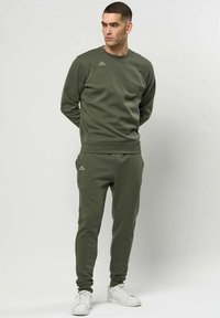 Mann stående med hendene bak ryggen, iført olivengrønn genser og joggebukser, samt hvite joggesko, på ensfarget lys bakgrunn.