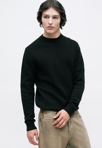 Νεαρός μεσαίου μήκους σκούρα μαλλιά, ντυμένος με έναν μαύρο πλεκτό πουλόβερ και μπεζ παντελόνι, που στέκεται μπροστά σε λευκό φόντο.