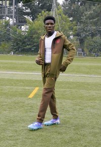 Survêtement de sport marron avec veste zippée et pantalon, T-shirt blanc en dessous, et chaussures de football colorées, marchant sur un terrain de gazon.