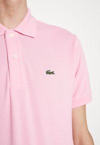 Camisa polo rosa hecha de tela texturizada, con un emblema de tres botones y un logo de cocodrilo verde en el lado izquierdo del pecho.