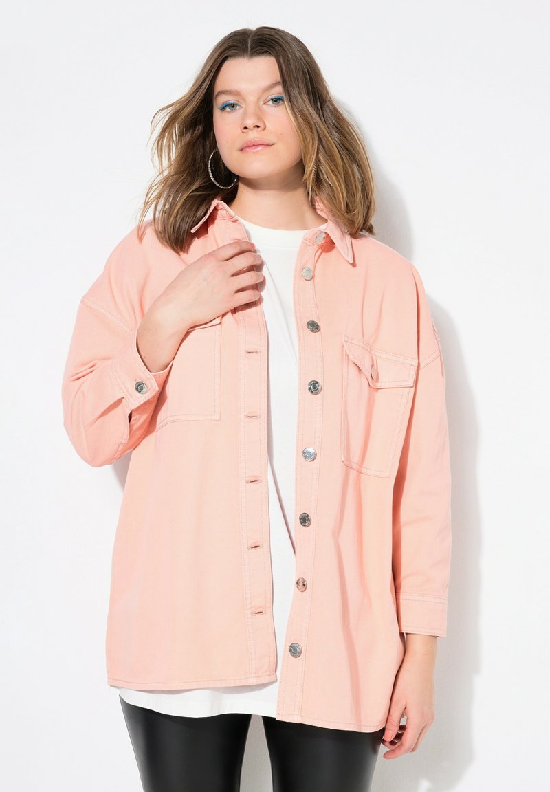 Camisa de mezclilla rosa claro con mangas largas, cierre de botones, bolsillo en el pecho y botones de metal, superpuesta sobre una camiseta blanca. Textura suave.