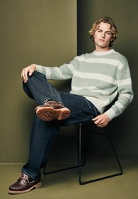 Brown leather boat shoes, dark denim jeans, green and white striped textured sweater, seated on a black wire chair against a neutral backdrop.