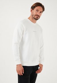 Biała bluza z miękkiego materiału, z okrągłym dekoltem, długimi rękawami i subtelnym haftowanym logo na piersi. Czarne dżinsy dopełniają stylizację.