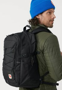 Black backpack with a sleek design, made of durable fabric, featuring front elastic cords, side pockets, and a logo patch. Wearer in a green jacket.