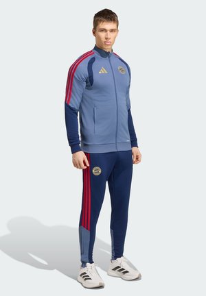 Młody mężczyzna stojący, ubrany w niebieski i granatowy dres Bayernu Monachium z czerwonymi paskami i białe buty adidas na jasnym tle.