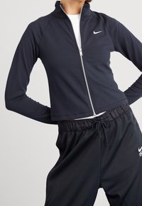 Svart zip-up jacka i slät tyg med hög krage. Har långa ärmar och en silverdragkedja, i kombination med svarta dragkedjebyxor.