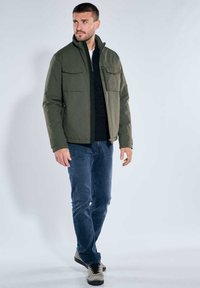 Olivgrüne Jacke mit Reißverschluss vorne, zwei Brusttaschen und elastischen Bündchen, kombiniert mit dunkelblauen Jeans und grauen Turnschuhen.