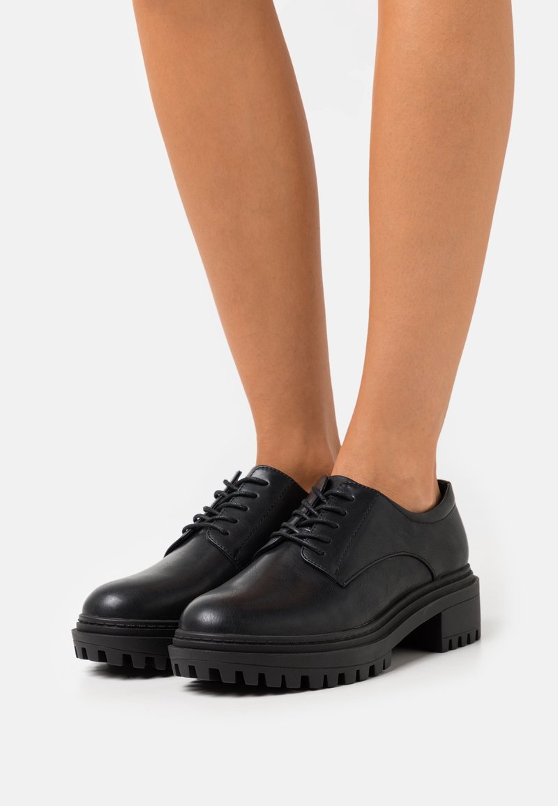 Chaussures en cuir noir à lacets avec semelles épaisses à crampons portées pieds nus sur un fond blanc uni.