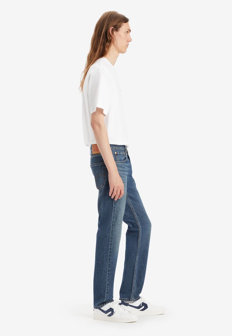 Leichtes weißes T-Shirt, kombiniert mit blauen Denim-Jeans; die Jeans haben einen geraden Schnitt, kontrastierende Nähte und ein Lederpatch an der Taille.