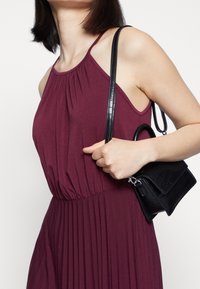 Kvinna klädd i en ärmlös burgundy klänning som bär en liten svart texturerad handväska med handtag och axelrem.