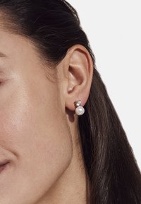 Zlatno-tonirane naušnice imaju okrugli biserni privjesak, osiguran s prozirnim kamenim akcentom na dnu. Glatka tekstura, jednostavan dizajn, elegantna završna obrada.