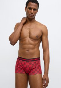 Κόκκινα ανδρικά μποξεράκια με μαύρη ελαστική μέση, διακοσμημένα με επαναλαμβανόμενο σχέδιο λογότυπου σε διάφορες αποχρώσεις του ροζ. Μαλακό, ελαστικό ύφασμα.