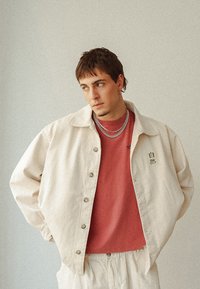 Beige knäppt jacka över en vinröd T-shirt, med en strukturerad tyg, två silverkedjor och en svart tryckt detalj på jackan.