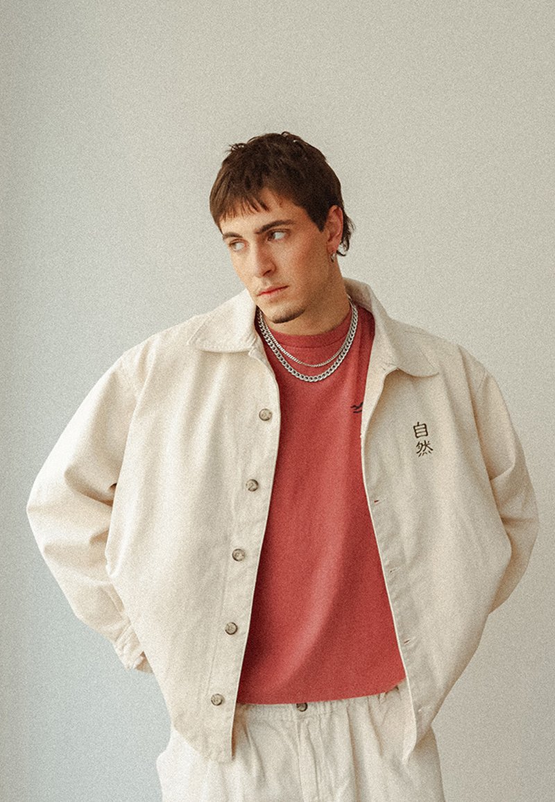 Beige knäppt jacka över en vinröd T-shirt, med en strukturerad tyg, två silverkedjor och en svart tryckt detalj på jackan.