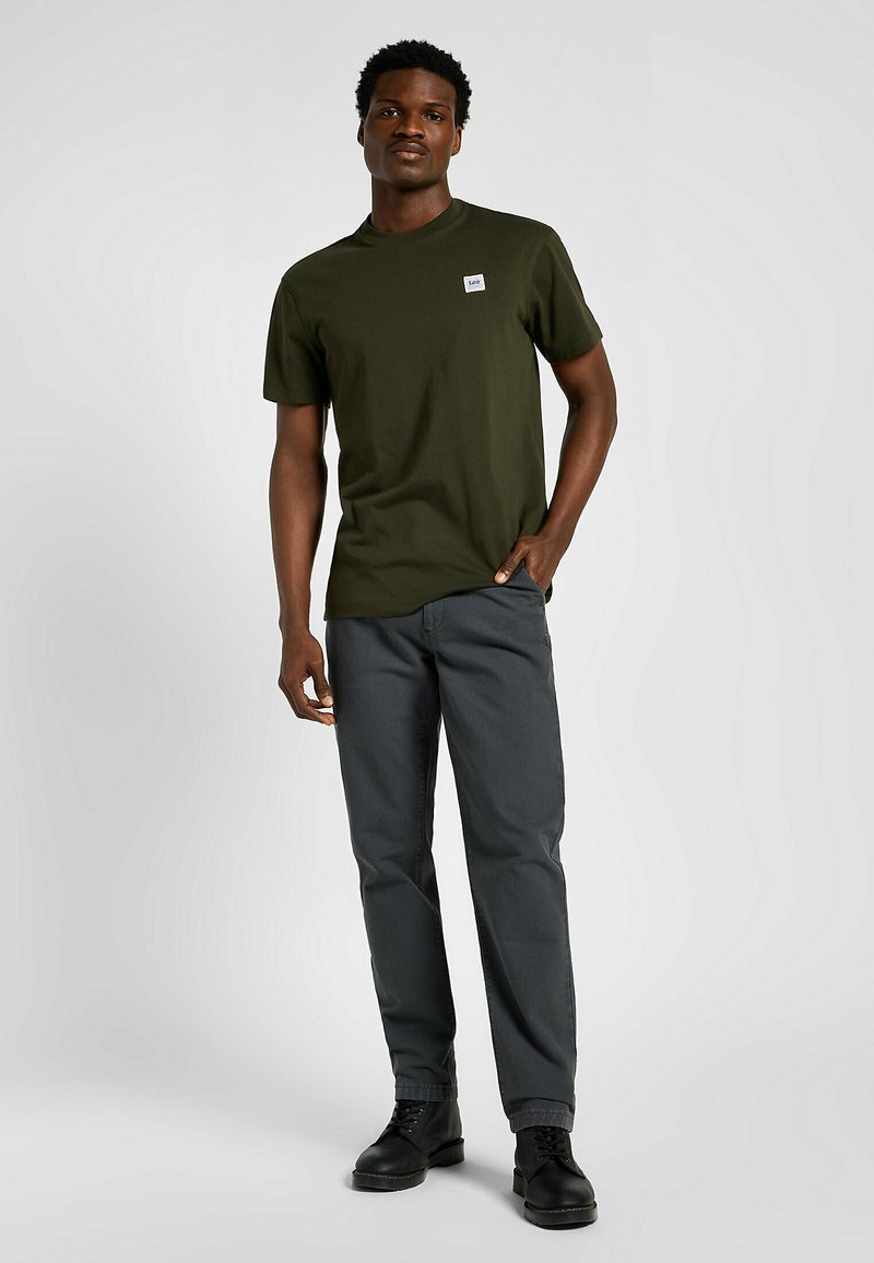 Oliwkowa koszulka z krótkim rękawem z małą łatą z logo, zestawiona z ciemnoszarymi spodniami i czarnymi butami na sznurówki, na neutralnym tle.