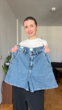 Νεαρή γυναίκα μέσα σε εσωτερικό χώρο κρατώντας ένα ζευγάρι υπερμεγέθη μπλε τζιν σορτς, χαμογελώντας απαλά, με φυτά και έπιπλα στο υπόβαθρο.