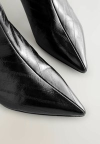 Decolletè nere a punta realizzate in materiale sintetico lucido, caratterizzate da una superficie testurizzata con linee diagonali e un tacco robusto.