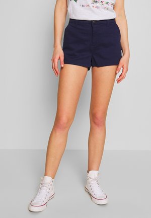 Frau trägt dunkelblaue Shorts, weiße hohe Sneaker und ein weißes T-Shirt mit Grafik und steht vor einem einfarbigen grauen Hintergrund.
