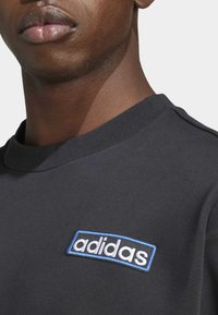 Černé bavlněné tričko s kulatým výstřihem a bílou nášivkou "adidas" s modrým okrajem na levé straně hrudi. Hladká textura, neformální design.