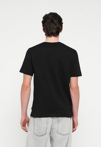 Μαύρο βαμβακερό μπλουζάκι, ορατό από πίσω, με κοντά μανίκια και στρογγυλή λαιμόκοψη. Σημαντική λεπτομέρεια: μικρό κόκκινο ταμπελάκι στο κάτω μέρος πλευράς.