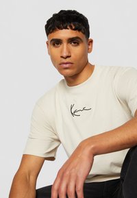 Beige cotton t-shirt with a round neck and short sleeves, featuring a black handwritten logo on the chest. Smooth texture, casual fit.