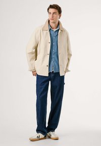 Joven con una chaqueta beige sobre una camisa azul de botones, jeans oscuros y zapatillas blancas con suelas negras y de goma.