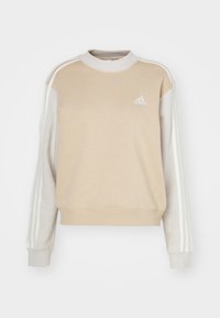 Béžová mikina s vysokým kulatým výstřihem, dlouhými rukávy a kontrastními světle béžovými bočními panely. Má tři bílé pruhy na každé paži a logo.