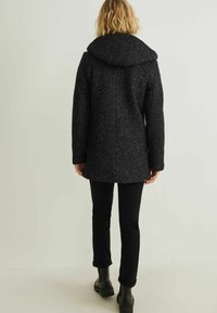 Persona vestida con un abrigo con capucha de color oscuro y moteado, pantalones negros y botas negras, de pie de espaldas frente a un fondo claro y liso.
