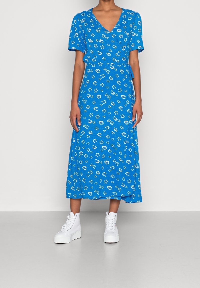 Blå omslagsklänning med korta ärmar och blommönster. Flynande tyg, knälång klippning, kombinerad med vita hög-top sneakers.