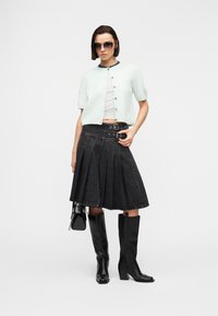 Γυναίκα που φοράει γυαλιά ηλίου, λευκή κοντομάνικη ζακέτα με κουμπιά, ριγωτό κοντό τοπ, μαύρη πλισέ μίντι φούστα με αγκράφες, μαύρες μπότες μέχρι το γόνατο και κρατάει τσάντα.