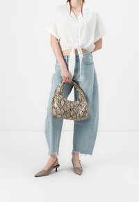 Donna che indossa una camicia bianca corta annodata, jeans azzurri con orlo sfrangiato, décolleté beige a punta con cinturino, mentre tiene una borsa in pitone beige e marrone.