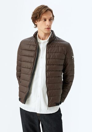 Chaqueta acolchada marrón con cremallera frontal, cuello alto y diseño acolchado horizontal. Detalles en negro en los puños y el dobladillo. Lleva una camisa blanca debajo.